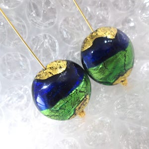 May include: A pair of handmade earrings featuring round, glass beads. The beads are a combination of blue, green, and gold leaf. Each bead is suspended from a thin, gold-colored wire. The earrings are displayed against a white background.