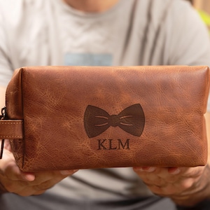 May include: Brown leather travel bag with a zipper closure and a bow tie design engraved on the front. The bag also has the initials KLM engraved on the front.