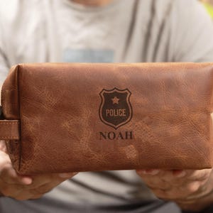 May include: Brown leather toiletry bag with a zipper closure and a leather handle. The bag has a personalized inscription that reads "Police" with a star inside a shield and "Noah" below it.