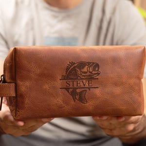 May include: A brown leather toiletry bag with a fish graphic and the name "STEVE" engraved on the front. The bag has a zipper closure and a small handle on the side. The bag is rectangular in shape.
