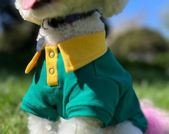 May include: A white dog wearing a green and yellow polo shirt. The dog is sitting in a grassy area.