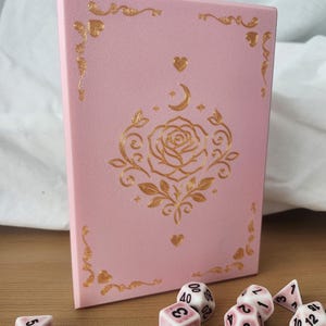 May include: A rectangular light pink box featuring a gold foil design of a central rose, a crescent moon, and small hearts. The box is accompanied by several polyhedral dice, each white with pink marbling and black numbers, scattered in front.