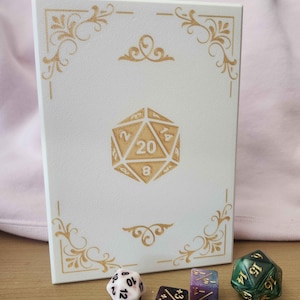 May include: A white rectangular box featuring gold filigree corner accents and a central gold 20-sided die graphic with the number 20 visible. Four polyhedral dice in various colors and shapes are positioned in front of the box on a flat surface.