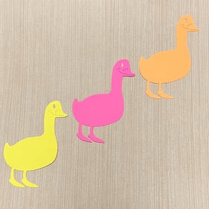 May include: Three paper duck cutouts in different colors: yellow, pink, and orange. The ducks are arranged in a row from left to right, with the yellow duck at the front, the pink duck in the middle, and the orange duck at the back.