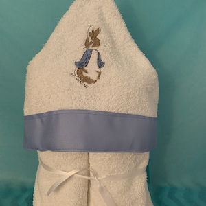 May include: White hooded towel with a blue ribbon trim and a Peter Rabbit embroidery. The towel is rolled up and tied with a white ribbon.
