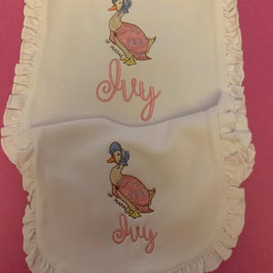 May include: White baby bibs with a ruffled edge, embroidered with a duck wearing a bonnet and the name "Ivy" in pink script. The duck is wearing a pink dress and blue bonnet. The bibs are on a pink background.