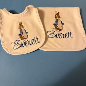 May include: White baby bib and blanket set. Each item features an embroidered image of a brown rabbit wearing a blue jacket. The name "Everett" is embroidered in blue script below the rabbit image. The bib has a curved shape, and the blanket is rectangular.