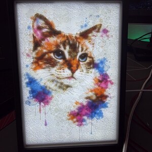 May include: A framed art print featuring a watercolor-style portrait of a cat. The cat has orange, brown, and white fur with blue eyes. Colorful paint splatters in blue, purple, and orange surround the cat's face, creating a vibrant and artistic effect.