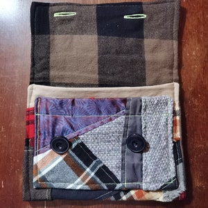 K&ouml;nnte beinhalten: Eine kleine, gepatchte Stofftasche mit einer braunen und schwarzen karierten Klappe. Die Tasche besteht aus verschiedenen Stoffen, darunter Karo, Blumen und einfarbige Stoffe. Sie hat zwei schwarze Kn&ouml;pfe und gr&uuml;ne N&auml;hte.