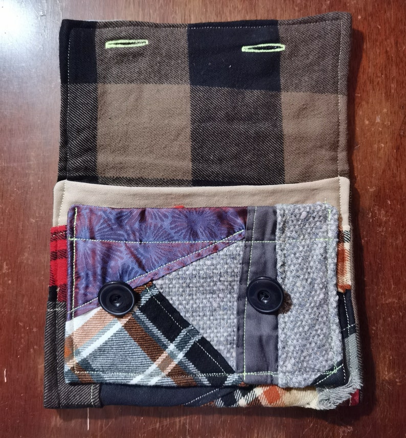 K&ouml;nnte beinhalten: Eine kleine, gepatchte Stofftasche mit einer braunen und schwarzen karierten Klappe. Die Tasche besteht aus verschiedenen Stoffen, darunter Karo, Blumen und einfarbige Stoffe. Sie hat zwei schwarze Kn&ouml;pfe und gr&uuml;ne N&auml;hte.