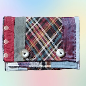 May include: A small patchwork wallet with a red, blue, and green plaid fabric, two white buttons, and a blue denim flap.