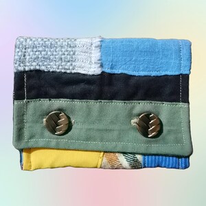 Op de afbeelding: Een multi-kleurige stoffen portemonnee met een patchwork ontwerp in geel, groen, blauw en zwart. De portemonnee heeft twee gouden knopen en een klep.