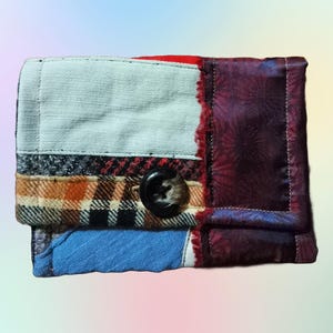 Op de afbeelding: Een klein patchwork portemonnee gemaakt van verschillende stoffen, waaronder ruit, denim en een bloemenprint. De portemonnee heeft een enkele knoopsluiting.