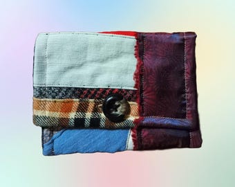 May include: A small patchwork wallet with a variety of fabrics, including plaid, denim, and a floral print. The wallet has a single button closure.