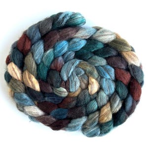 May include: A thick, braided coil of roving wool fiber featuring a variegated color palette of deep forest green, navy blue, earthy brown, muted teal, and soft beige tones, arranged in a circular spiral pattern with a soft, fuzzy texture.