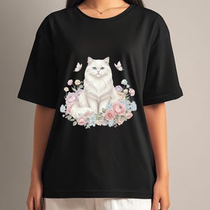May include: Black t-shirt with a white cat illustration, blue eyes, and a floral wreath of pink, blue, and white flowers. Two butterflies are on either side of the cat. The shirt has short sleeves.