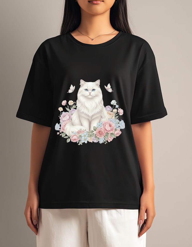 May include: Black t-shirt with a white cat illustration, blue eyes, and a floral wreath of pink, blue, and white flowers. Two butterflies are on either side of the cat. The shirt has short sleeves.