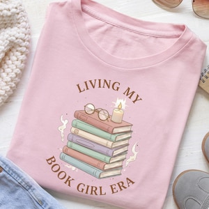 May include: A pale pink t-shirt featuring the phrase "LIVING MY BOOK GIRL ERA" encircling a graphic of books, spectacles, and a candle. The books are in different colours, and the design has an illustrated style.