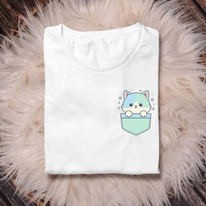 K&ouml;nnte beinhalten: Wei&szlig;es T-Shirt mit einer Tasche, die eine Cartoon-Katze mit blauen und t&uuml;rkisfarbenen Akzenten zeigt. Die Katze schaut aus einer mintgr&uuml;nen Tasche, umgeben von kleinen Sternen. Das Shirt ist auf einer flauschigen, wei&szlig;en Oberfl&auml;che gefaltet.