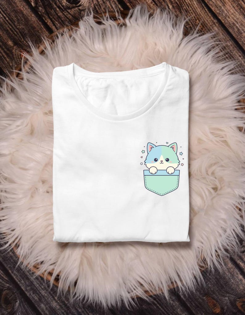 K&ouml;nnte beinhalten: Wei&szlig;es T-Shirt mit einer Tasche, die eine Cartoon-Katze mit blauen und t&uuml;rkisfarbenen Akzenten zeigt. Die Katze schaut aus einer mintgr&uuml;nen Tasche, umgeben von kleinen Sternen. Das Shirt ist auf einer flauschigen, wei&szlig;en Oberfl&auml;che gefaltet.