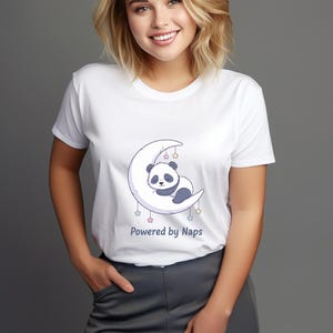Może przedstawiać: Biała koszulka z nadrukiem przedstawiającym śpiącą pandę na księżycu w kształcie półksiężyca, ozdobionym gwiazdami. Pod grafiką znajduje się napis "Powered by Naps". Koszulka jest w stylu casual.
