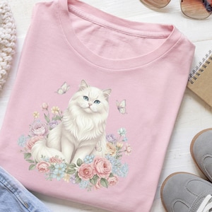 May include: A pale pink crew neck t-shirt with a watercolour design of a white cat with blue eyes, flowers, and butterflies. The cat is the central image, with a fluffy appearance. The design is in a soft, watercolour style.