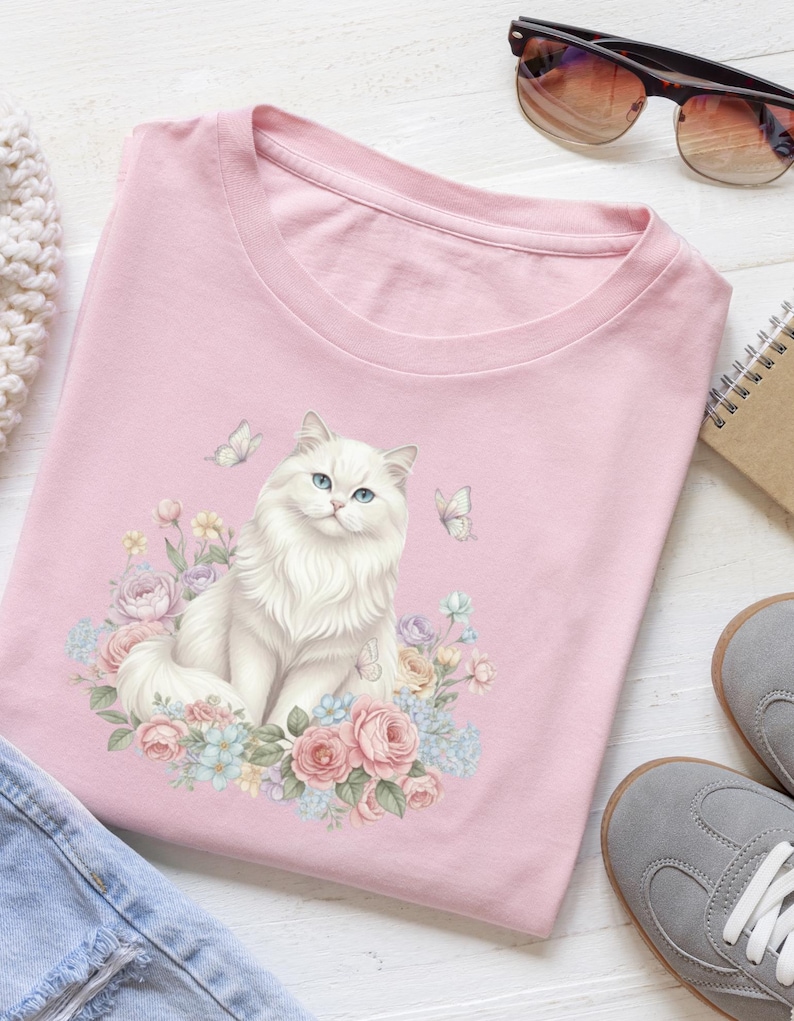 May include: A pale pink crew neck t-shirt with a watercolour design of a white cat with blue eyes, flowers, and butterflies. The cat is the central image, with a fluffy appearance. The design is in a soft, watercolour style.
