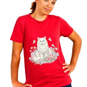 May include: A red t-shirt with a graphic of a white cat with blue eyes, encircled by pastel flowers and butterflies. The design is centered on the shirt, with a bright red backdrop.