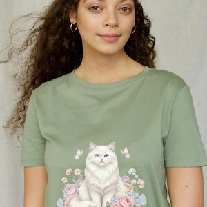 May include: Sage green t-shirt with a white cat illustration. The cat has blue eyes and is surrounded by pastel flowers and butterflies. The t-shirt is a crew neck style.