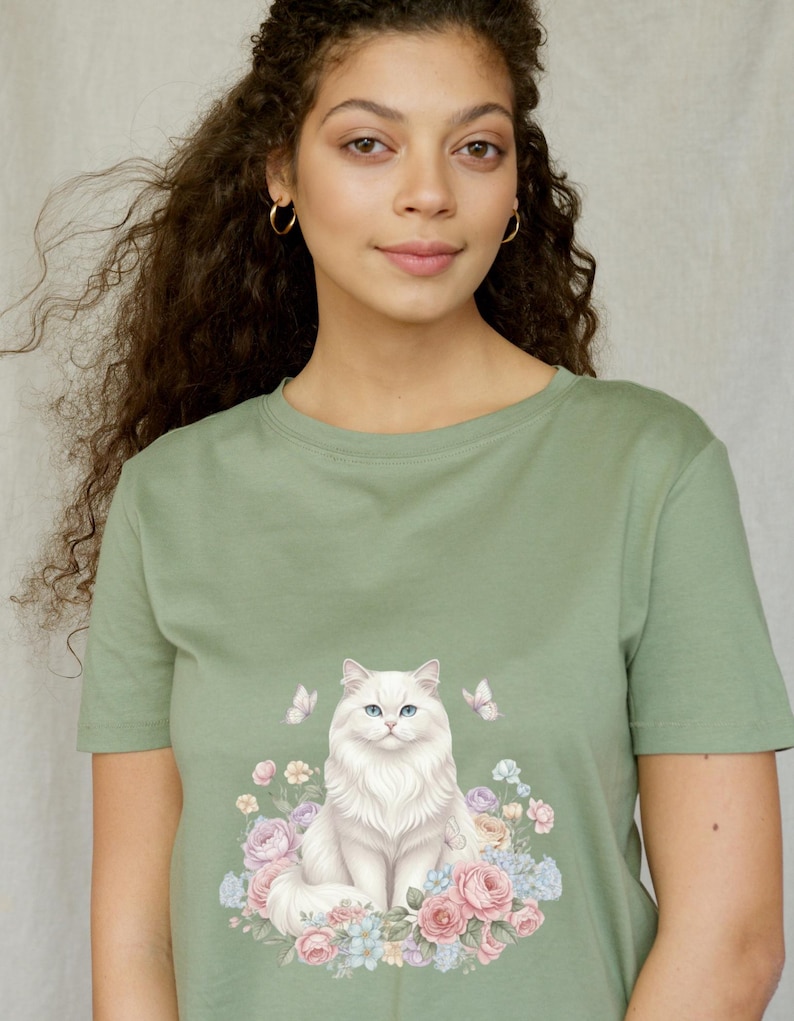 May include: Sage green t-shirt with a white cat illustration. The cat has blue eyes and is surrounded by pastel flowers and butterflies. The t-shirt is a crew neck style.