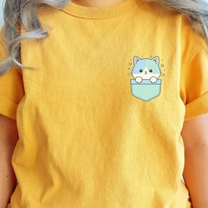 K&ouml;nnte beinhalten: Senfgelbes T-Shirt mit einer hellblauen Tasche mit einem Cartoon-Katzen-Design. Die Katze hat blaues und wei&szlig;es Fell und ist von kleinen Sternen umgeben. Das Shirt hat kurze &Auml;rmel und einen Rundhalsausschnitt.