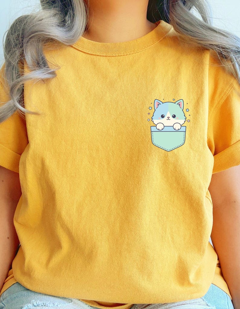 K&ouml;nnte beinhalten: Senfgelbes T-Shirt mit einer hellblauen Tasche mit einem Cartoon-Katzen-Design. Die Katze hat blaues und wei&szlig;es Fell und ist von kleinen Sternen umgeben. Das Shirt hat kurze &Auml;rmel und einen Rundhalsausschnitt.