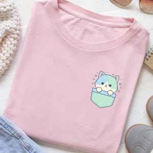 K&ouml;nnte beinhalten: Hellrosa T-Shirt mit einem Cartoon-Katzenmotiv, das aus einer Tasche schaut. Die Katze hat blaue und gr&uuml;ne Akzente, und die Tasche ist gr&uuml;n umrandet. Kleine Sterne umgeben die Katze. Das T-Shirt liegt gefaltet auf einer wei&szlig;en Holzoberfl&auml;che.