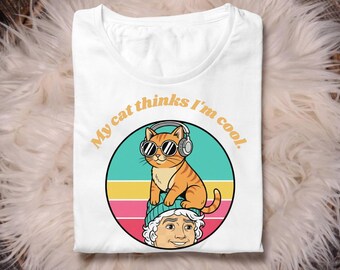 May include: White t-shirt with a graphic of an orange tabby cat wearing sunglasses and headphones, sitting on a person's head. The text "My cat thinks I'm cool" is arched above the cat. The design is set against a retro striped background.