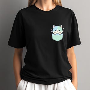 K&ouml;nnte beinhalten: Schwarzes T-Shirt mit einer Cartoon-Katze, die aus einer Tasche schaut. Die Katze hat blaues und wei&szlig;es Fell mit Sternen. Die Tasche ist hellgr&uuml;n. Das T-Shirt hat einen l&auml;ssigen Stil.