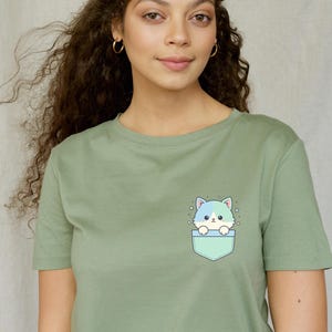 K&ouml;nnte beinhalten: Salbeigr&uuml;nes T-Shirt mit einem Cartoon-Katzen-Design. Die Katze mit blau-wei&szlig;em Fell befindet sich in einer hellgr&uuml;nen Tasche. Das T-Shirt hat einen Rundhalsausschnitt.