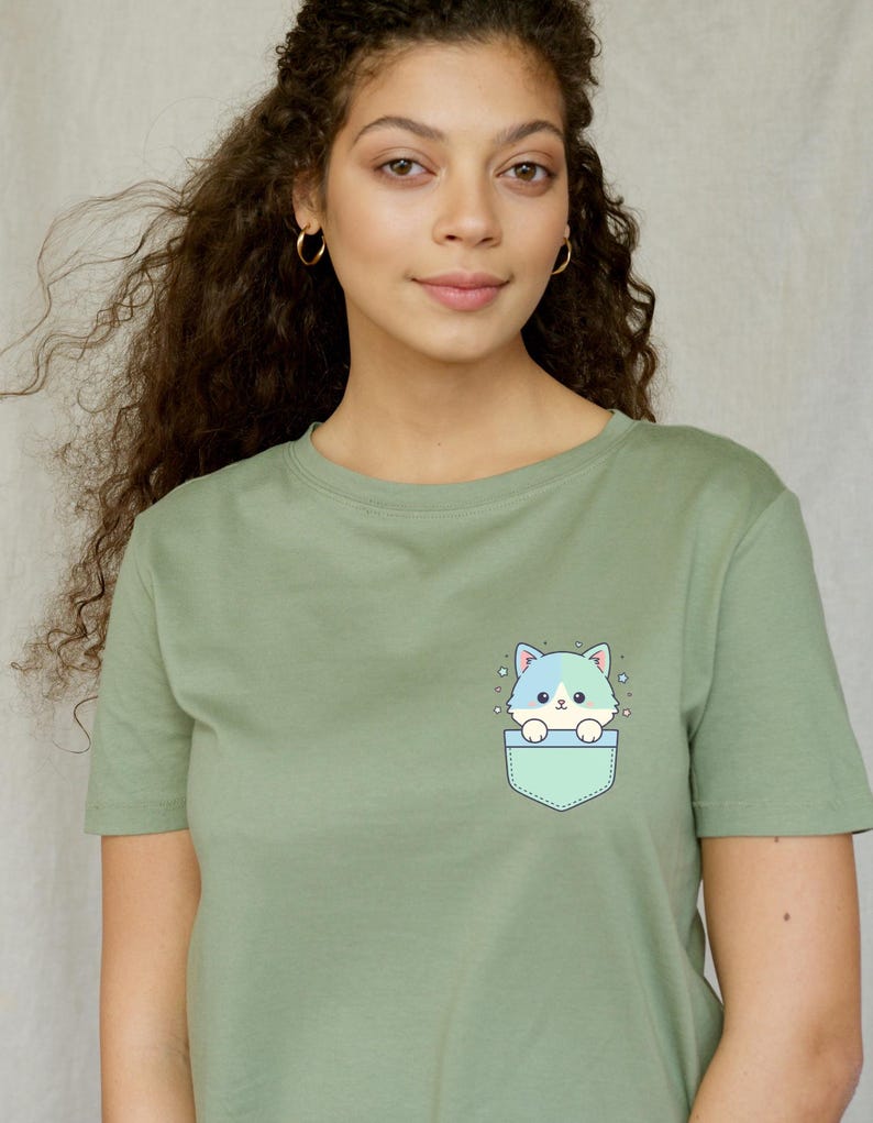K&ouml;nnte beinhalten: Salbeigr&uuml;nes T-Shirt mit einem Cartoon-Katzen-Design. Die Katze mit blau-wei&szlig;em Fell befindet sich in einer hellgr&uuml;nen Tasche. Das T-Shirt hat einen Rundhalsausschnitt.