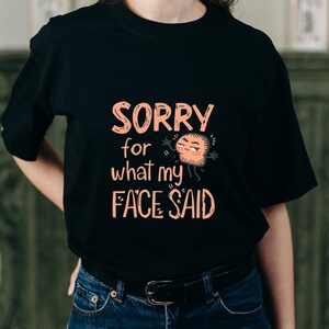 Puede incluir: Camiseta negra con el texto "SORRY for what my FACE SAID" en naranja. La camiseta presenta un gráfico de cara de dibujos animados con brazos. La persona lleva vaqueros azules y un cinturón negro.