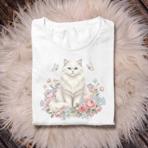 May include: White t-shirt with a detailed illustration of a fluffy white cat with blue eyes, surrounded by pastel-coloured flowers and butterflies. The design is centered on the shirt, creating a whimsical and artistic aesthetic. The shirt is folded neatly.
