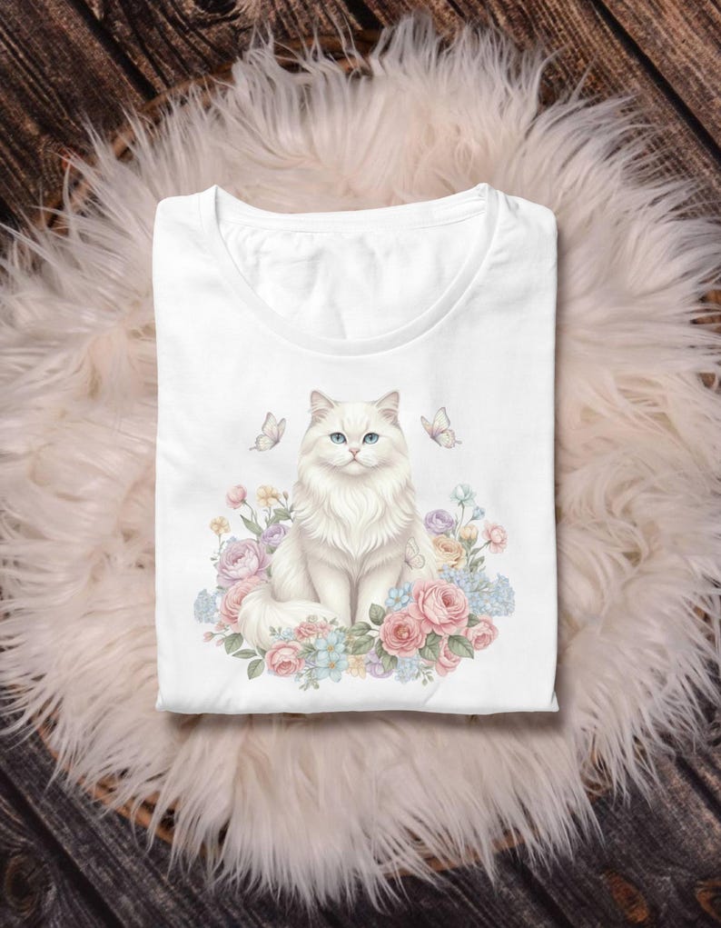 May include: White t-shirt with a detailed illustration of a fluffy white cat with blue eyes, surrounded by pastel-coloured flowers and butterflies. The design is centered on the shirt, creating a whimsical and artistic aesthetic. The shirt is folded neatly.