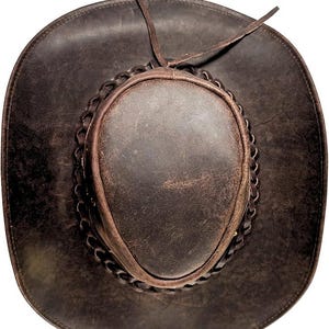 Pode incluir: Chap&eacute;u de cowboy de couro castanho escuro com aba larga e faixa tran&ccedil;ada ao redor da coroa. O chap&eacute;u tem um acabamento desgastado e uma gravata de couro. Este chap&eacute;u de estilo ocidental &eacute; perfeito para atividades ao ar livre.