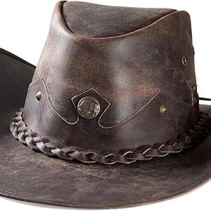 Pode incluir: Chap&eacute;u de cowboy de couro castanho desgastado com uma faixa tran&ccedil;ada e detalhes met&aacute;licos decorativos. O chap&eacute;u apresenta uma aba larga e um design ocidental cl&aacute;ssico, ideal para atividades ao ar livre ou eventos &agrave; fantasia.