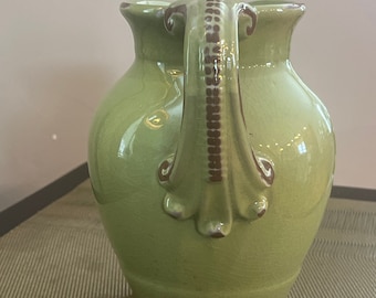 May include: A green ceramic vase with a decorative handle. The vase has a glossy finish and a slightly rounded shape.