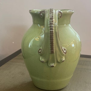 Peut inclure: Un vase en céramique vert avec une poignée décorative. Le vase a une finition brillante et une forme légèrement arrondie.