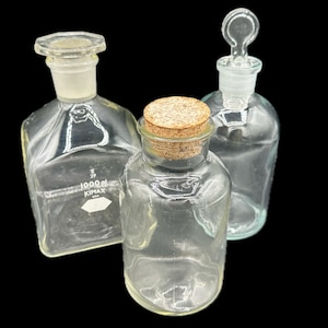 May include: Three clear glass laboratory bottles. One bottle has a cork stopper, another has a glass stopper, and the third has a glass stopper. One bottle is labeled "1000 ml".