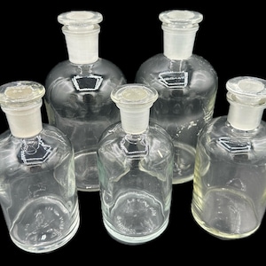 May include: Five clear glass bottles with glass stoppers. The bottles are of varying sizes and shapes, with rounded bodies and flared necks. The stoppers are a frosted glass. The bottles are arranged against a black background.