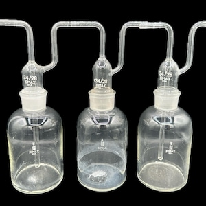 May include: Three clear glass laboratory flasks with curved tubes and ground glass joints. Each flask has a round base and a bulbous body. The flasks are labeled with "34/28" and "KIMAX USA".