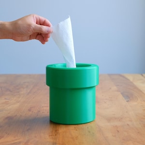 May include: A cylindrical green tissue holder with a wide, flat rim at the top. A single white tissue is being pulled upwards from the centre opening of the container. The object has a smooth, solid-coloured surface and a distinct pipe-like shape.