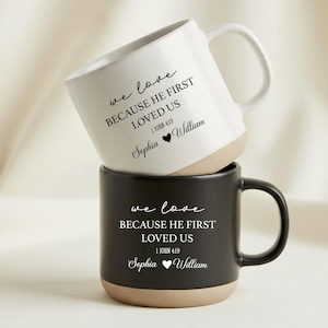 May include: Two stacked ceramic mugs. The top mug is white with a speckled finish and a white handle. The bottom mug is black. Both mugs have the text "we love BECAUSE HE FIRST LOVED US 1 JOHN 4:19 Sophia William" printed in black.