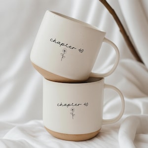 Può includere: Due tazze in ceramica bianca con base beige sono impilate. Ogni tazza ha le parole "chapter 40" e una piccola illustrazione floreale stampata in nero. Le tazze hanno un design semplice e moderno.
